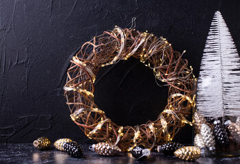 White, black New Year decorations. Handmade wreath with fairy lights  and  jar with decorative white tree and glass cones against black textured background. Scandinavian minimalistic style. Still life
