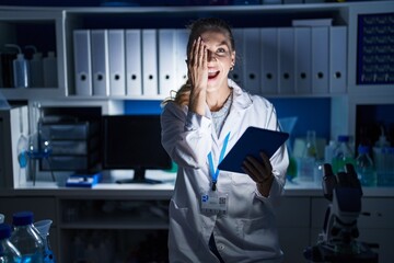 Beautiful blonde woman working at scientist laboratory late at night covering one eye with hand, confident smile on face and surprise emotion.