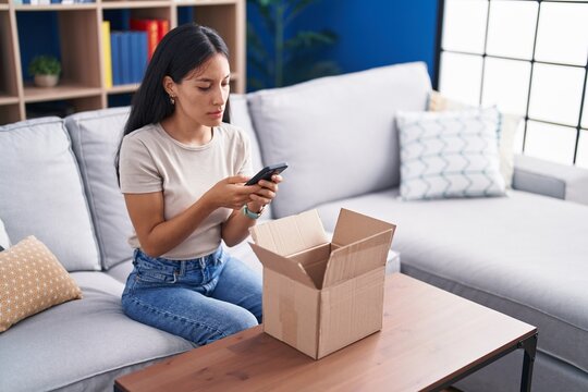 Young beautiful hispanic woman using smartphone unpacking cardboard box at home