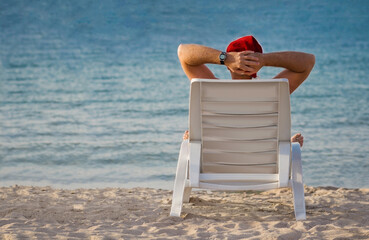 A man in a Santa hat is sitting in sun loungers by the seashore with his hands behind his head, looking at the water and relaxing on vacation. Santa is on vacation at the beach.