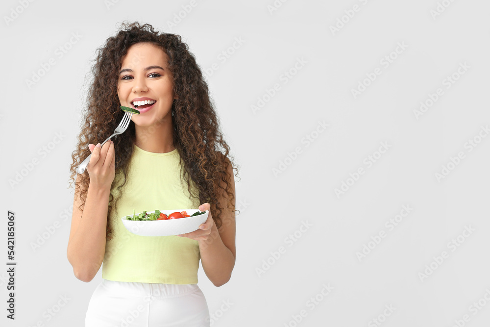 Wall mural Happy young African-American woman with fresh salad on light background. Diet concept