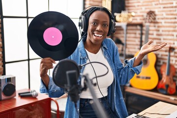 Beautiful black woman holding vinyl record at music studio celebrating achievement with happy smile and winner expression with raised hand