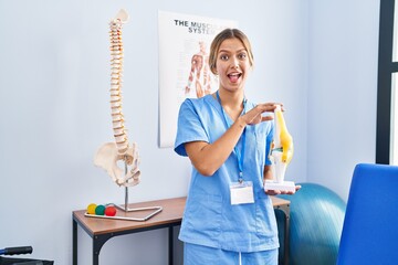 Young blonde orthopedic woman holding anatomical model of knee joint celebrating crazy and amazed...