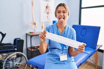 Young blonde woman holding therapy strip band sticking tongue out happy with funny expression.