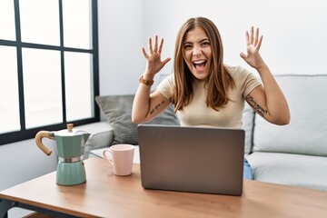 Young brunette woman using laptop at home drinking a cup of coffee celebrating crazy and amazed for success with arms raised and open eyes screaming excited. winner concept