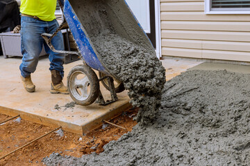 On side house construction worker pours cement from wheelbarrow to create concrete sidewalk that...