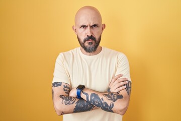 Hispanic man with tattoos standing over yellow background skeptic and nervous, disapproving expression on face with crossed arms. negative person.