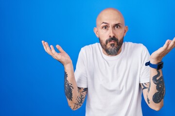 Hispanic man with tattoos standing over blue background clueless and confused expression with arms...