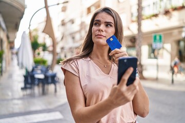 Young beautiful hispanic woman using smartphone and credit card with doubt expression at street