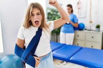 Blonde little girl wearing arm on sling at rehabilitation clinic annoyed and frustrated shouting...