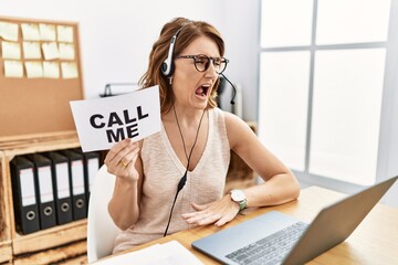 Middle age brunette woman wearing operator headset holding call me banner angry and mad screaming...