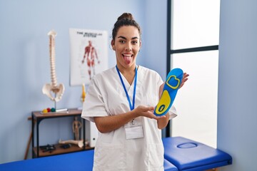 Young hispanic woman holding shoe insole at physiotherapy clinic sticking tongue out happy with funny expression.