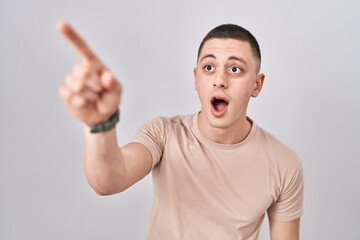 Young man standing over isolated background pointing with finger surprised ahead, open mouth amazed expression, something on the front