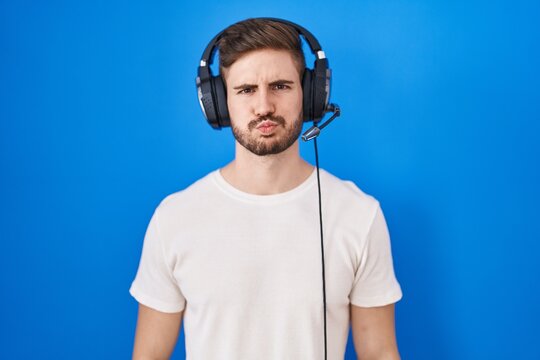 Hispanic man with beard listening to music wearing headphones puffing cheeks with funny face. mouth inflated with air, crazy expression.