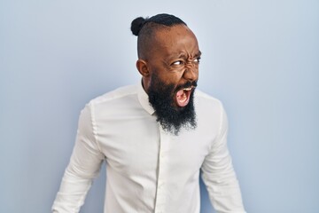 African american man standing over blue background angry and mad screaming frustrated and furious,...