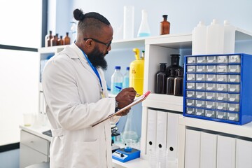 Naklejka premium Young african american man wearing scientist uniform writing on clipboard at laboratory