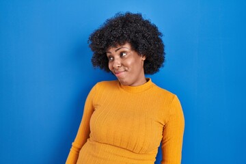 Fototapeta na wymiar Black woman with curly hair standing over blue background smiling looking to the side and staring away thinking.