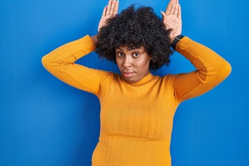 Fototapeta na wymiar Black woman with curly hair standing over blue background doing bunny ears gesture with hands palms looking cynical and skeptical. easter rabbit concept.