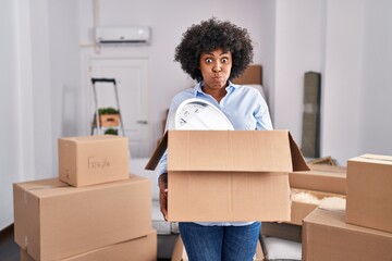 Black woman with curly hair moving to a new home holding cardboard box puffing cheeks with funny face. mouth inflated with air, catching air. - obrazy, fototapety, plakaty
