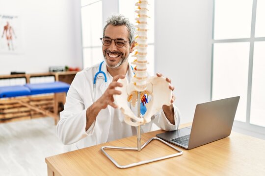 Middle age grey-haired man doctor holding anatomical model of spinal column at rehab clinic