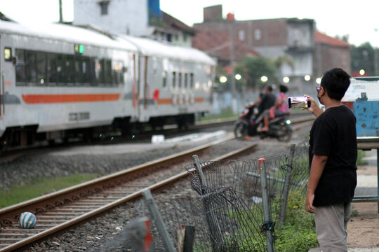 A boy captures with his cellphone a scooter rider breaking a railroad crossing when a train passes. Selective focus.