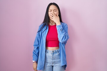 Young asian woman standing over pink background bored yawning tired covering mouth with hand. restless and sleepiness.