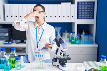 Young brunette woman working at scientist laboratory peeking in shock covering face and eyes with hand, looking through fingers with embarrassed expression.