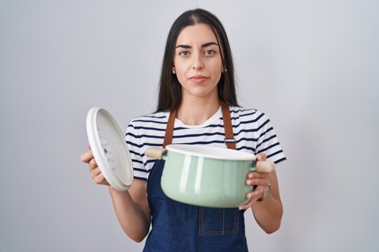 Young brunette woman wearing apron holding cooking pot relaxed with serious expression on face. simple and natural looking at the camera.