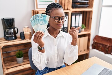 African woman with braids working at the office holding brazilian reals pointing thumb up to the side smiling happy with open mouth