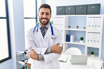 Handsome hispanic man wearing doctor uniform and stethoscope at medical clinic happy face smiling with crossed arms looking at the camera. positive person.