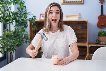 Blonde caucasian woman holding piggy bank and hammer celebrating crazy and amazed for success with open eyes screaming excited.