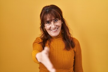 Middle age hispanic woman standing over yellow background smiling friendly offering handshake as greeting and welcoming. successful business.