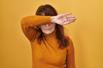 Middle age hispanic woman standing over yellow background covering eyes with arm, looking serious...