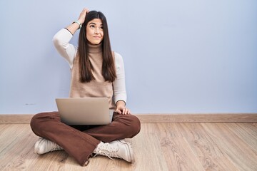 Young brunette woman working using computer laptop sitting on the floor confuse and wondering about...