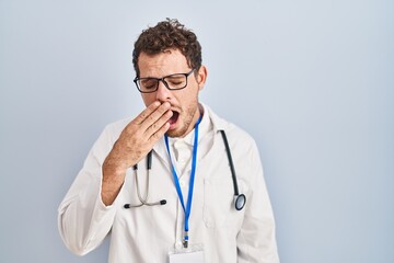 Young hispanic man wearing doctor uniform and stethoscope bored yawning tired covering mouth with...