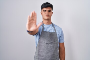 Hispanic young man wearing apron over white background doing stop sing with palm of the hand. warning expression with negative and serious gesture on the face.