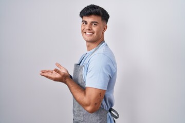 Hispanic young man wearing apron over white background pointing aside with hands open palms showing copy space, presenting advertisement smiling excited happy