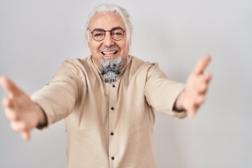 Middle age man with grey hair standing over isolated background looking at the camera smiling with open arms for hug. cheerful expression embracing happiness.