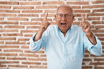 Senior man with grey hair standing over bricks wall smiling amazed and surprised and pointing up with fingers and raised arms.