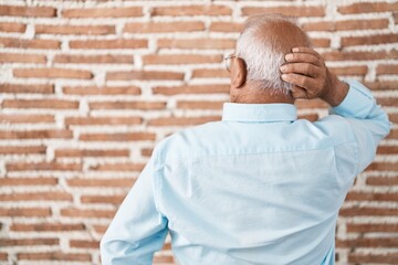 Senior man with grey hair standing over bricks wall backwards thinking about doubt with hand on head