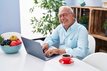 Senior grey-haired man using laptop and drinking coffee sitting on table at home