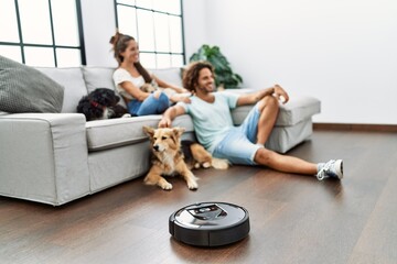 Man and woman couple sitting on sofa with dogs cleaning floor using vacuum robot at home - obrazy, fototapety, plakaty