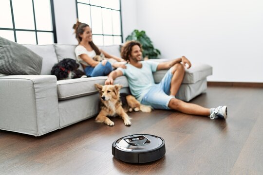 Man and woman couple sitting on sofa with dogs cleaning floor using vacuum robot at home