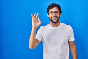 Fototapeta premium Handsome latin man standing over blue background smiling positive doing ok sign with hand and fingers. successful expression.
