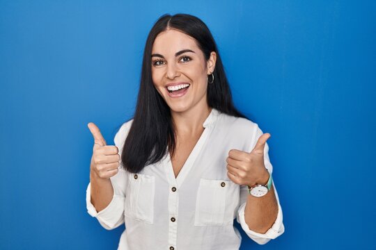 Young hispanic woman standing over blue background success sign doing positive gesture with hand, thumbs up smiling and happy. cheerful expression and winner gesture.