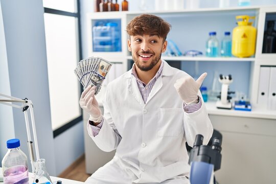 Arab man with beard working at scientist laboratory holding money pointing thumb up to the side smiling happy with open mouth