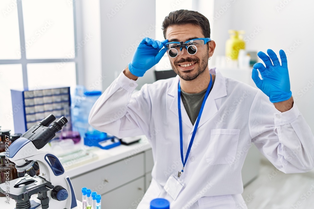 Wall mural Young hispanic man working at scientist laboratory wearing magnifying glasses doing ok sign with fingers, smiling friendly gesturing excellent symbol