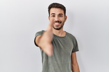 Young hispanic man with beard wearing casual t shirt over white background smiling friendly offering handshake as greeting and welcoming. successful business.