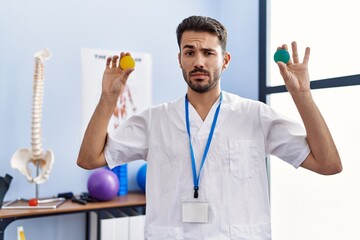 Young hispanic physiotherapist man holding strength balls to train hand muscles skeptic and nervous, frowning upset because of problem. negative person.