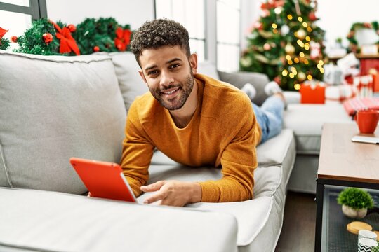 Young arab man using touchpad lying on the sofa by christmas tree at home.